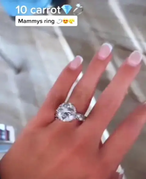 TikTok A large diamond ring set sits on a woman's finger, as she has her hand outstretched. It's set in silver, and has smaller diamonds running down the band of the ring. The background of the image is blurred. Above some text says "10 carrot" followed by a diamond emoji and a ring emoji. Below that is a white text box which reads "mammy's ring" followed by another ring emoji and a heart eye emoji. 