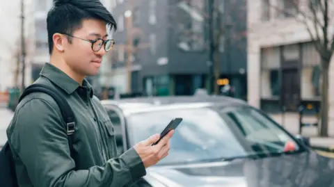 Man holding a phone stands next to a black car in a street.