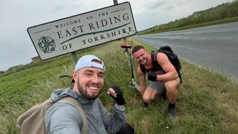 Joel Redhead Two men clenching their fists as they crouch next to a sign at the side of a road that reads "Welcome to the East Riding of Yorkshire". One man is wearing a white cap backwards, a grey coat and a beige backpack. The other man is wearing a black vest, a backpack and grey shorts.
