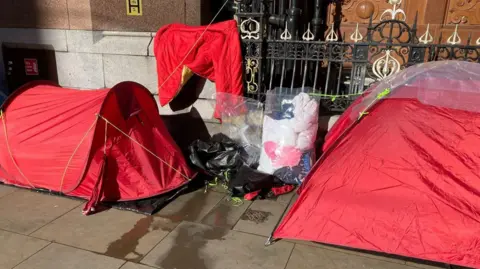 Two red tents outside Mosley Street in Manchester city centre. A red sleeping bag can be seen hung up on a rail, with a plastic, transparent bag seen containing clothes propped up on a rail fence between the two tents. 