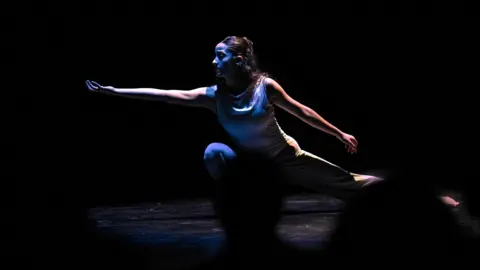 a contemporary dancer onstage, reaching with her hand turned towards the ceiling; one of her legs is in a deep squat (stock image)