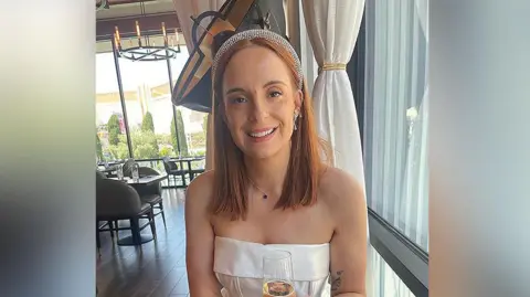 Cancer Research UK handout Liv Shaw with long auburn hair wearing a diamante tiara and white silk corset style wedding dress. She is in a restaurant holding a glass of champagne to celebrate her wedding and smiling.