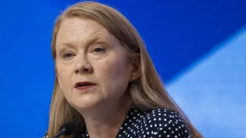 A woman with light-coloured hair sits on a stage in front of a blue background. She is visible from the shoulders up and is wearing a polka dot black and white shirt. 