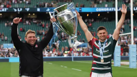 Action Images/Reuters Two smiling men standing with their arms aloft holding a large silver trophy between them. The man on the left has blond hair and is wearing a black top, the other is wearing a Leicester Tigers green and red rugby kit and a winners medal. They are standing on a rugby pitch with a fairly empty stand behind them.