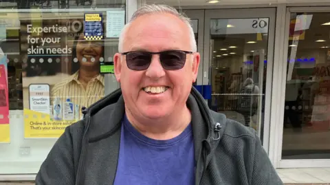 Henry Godfrey-Evans/BBC Keith Horner has white hair, darkly tinted glasses, a blue t-shirt and a darkly-coloured hooded top. He is sitting or standing in the high street, and a shop frontage is behind him.