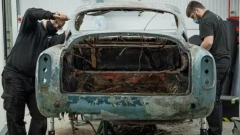 Marc Bow/Timbuktu Content Two technicians stand either side of the damaged chassis of the Aston Martin as they begin to repair it in a workshop