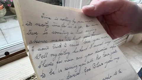 Mike Gould A letter being held up. The part we can see reads:
Time is rolling by, once more it's nearly time for us to go. I'm sure there is something in that saying absence makes the heart grow fonder. I don't think I could love you more. Well, my darling husband I must say cheerio for now and your devoted wife to be sending you all her love.