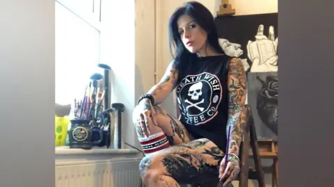 A heavily-tattooed woman with black hair is sitting on a stool in an art studio and posing for a picture. There are paintbrushes on a windowsill and there is a portrait on the wall behind her.