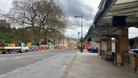 An image of Queen Street with a road closure in place. The entrance to York Station is visible to the right, with York's walls on the left at the far side of the road.