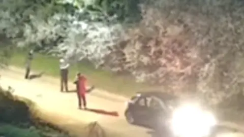 Suffolk Police Grainy CCTV footage showing three figures standing in a road while seemingly looking into trees. On the right of the image is a dark coloured car with its lights on.