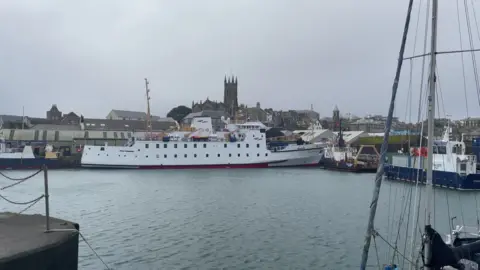 The white vessel sits alongside in Penzance harbour. The town and a church spire are visible behind her. 