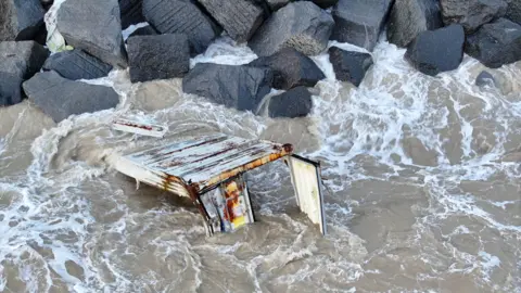 A rusty, white shipping container floating in the sea. It is extensively damaged, missing its top and bottom and with a broken door. It is mostly submerged in water, and appears stuck on a wall of large black rocks.