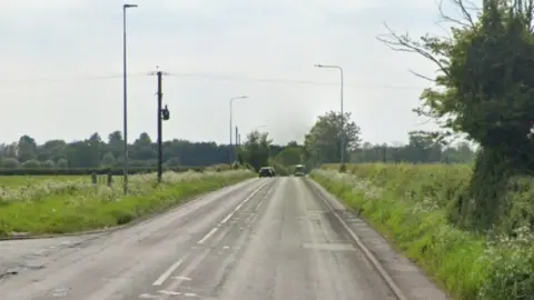 A main road, with a junction to the left, and fields and trees on either side
