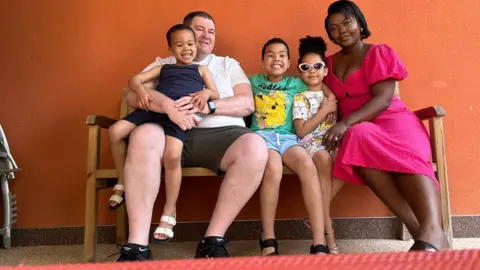 Adrian Scarlett Adrian Scarlett sits on a bench, in front of an orange wall, with his wife and three children.