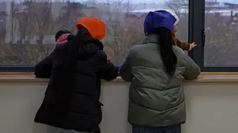 Dos mujeres están de pie frente a una ventana del Gurdwara. Llevan abrigos acolchados y pañuelos en la cabeza y sostienen a niños pequeños.