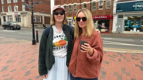 Lili Sheppard/BBC A woman with a pink cap, grey cardigan and white shirt and pale blue dress. She is standing next to a blonde woman with a rusty-coloured coat and holding a cup of coffee. They are standing on a high street.