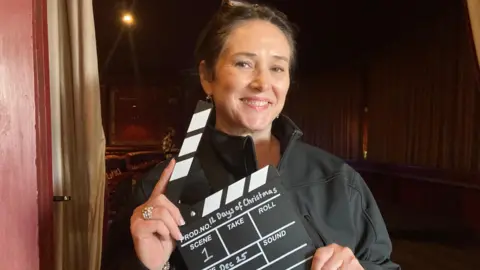 Lynn Dockerty Lynn Dockerty has swept back light brown hair and is holding a clapper board with 12 Days Of Christmas written on it.