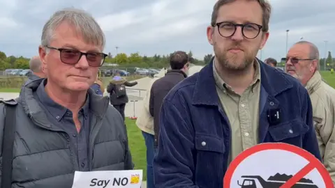 Lea Park Residents Association Henley and Thame MP Freddie Van Mierlo attending the demonstration, holding a sign that had a lorry crossed out. Another resident is next to him, holding a sheet of paper that says "Says NO". Other people could be seen behind them. It's an overcast day.