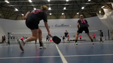 Jamie Niblock/BBC A female player stoops down to reach a shot. She is about to make connection with the luminous orange polymer ball, while her doubles partner is poised for the ball to be returned. In the background, their opponents are on the other side of the net. 