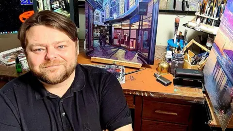 A man with brown hair and a beard looking at the camera. He is sitting in front of a painting which sits on a desk beside paint brushes, sprays and painting colours.