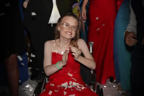 Chloe Russell A woman wearing a bright red ballgown smiles as she is covered in paper confetti. She is surrounded by other people wearing formal clothing.