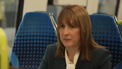 Rachel Reeves, a woman with shoulder length brown hair and brown eyes. She is wearing a cream blouse and navy suit jacket. She is being interviewed in a train carriage