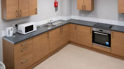 Shropshire Council A kitchen with a pale floor, wooden units and a black worktop. There is an oven, a kettle and a microwave and three wall-mounted units