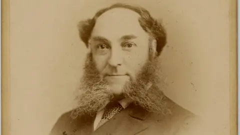 John Fraser archive Sepia photo of William Whiteley, 1907. A balding man wearing a suit in a formal portrait. He has massive whiskers on each side of his chin .