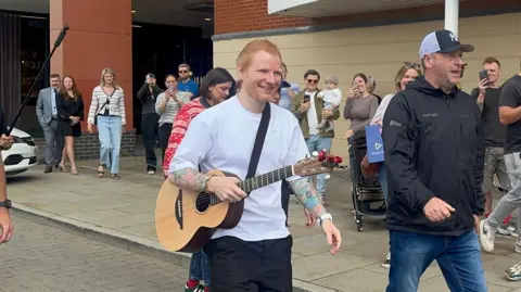 A man with red hair and a white T-shirt walks along a road with a guitar strapped around his torso. Crowds of people take photos of him from the sides of the road.