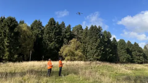 Image shows drone operators standing in a field looking up at the drone in the air. They both wear orange jackets. 