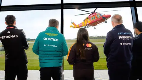 Representatives from Formula 1 are seen with their backs to camera looking out of big windows at a Cornwall Air Ambulance helicopter 
