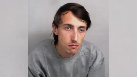 Essex Police A police custody photo of Charlie Land, who wears a grey jumper and is positioned in front of a grey backdrop. He has long dark hair in a side parting and stubble