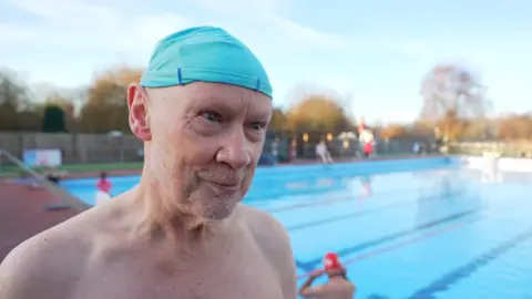 Alex Dunlop/BBC A head and shoulders image of Nigel Tebb. He is wearing a light blue swimming cap and looking past the camera. He is topless and standing around the side of the pool. 