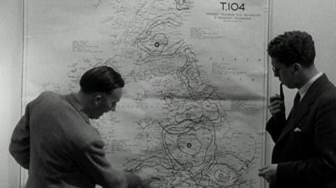 A man gesturing at locations on a map of the UK on a wall.