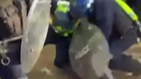 Supplied Two police officers are wearing riot gear and are on top of a man on the ground.