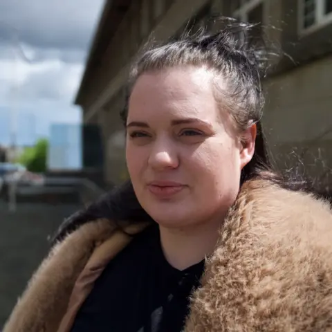 Fiona, a woman with dark hair partly tied back, wearing a jacket with a fur collar - she is outside with a large building behind her