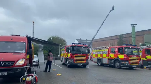 A photograph from the scene that shows three fire engines, a fire service command unit van and an aerial ladder platform. There are clouds of grey smoke in the sky above 