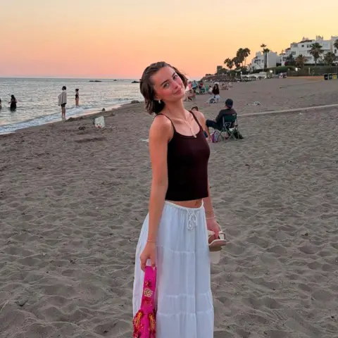 Erin Lavery Person standing on a sandy beach at sunset, wearing a dark sleeveless top and a white skirt, holding sandals in one hand and a bright pink accessory in the other. The sea stretches along the shoreline, with several people near the water and white buildings and palm trees visible in the distance beneath a pastel orange and pink sky.