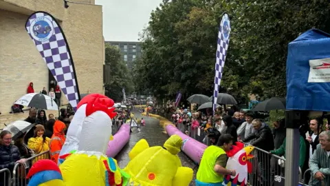 A racetrack going downhill with spectators on both sides behind railings. Some of them have umbrellas up. Two purple and white chequered banners are flying. In the foreground are people dressed in inflatable chicken outfits.