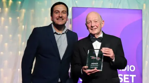 Andy Bennett wearing a dark suit and blue-grey shirt standing beside Gordon Swindells, who is wearing a black suit and a bow tie, holding up his glass award and smiling at the camera. There is a purple Radio Somerset banner screen behind them.