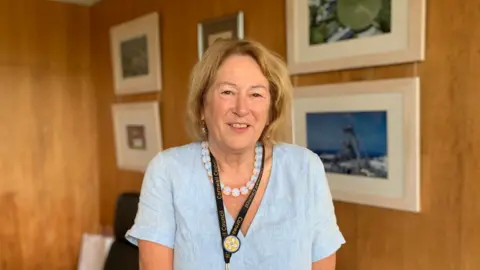 BBC Linda Taylor wearing a light blue v-necked top with a beaded necklace and a black Cornwall Council lanyard. She has short, shoulder-length hair and is smiling at the camera.