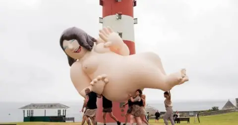 A giant model of a naked woman is carried by a group of people past a red and white striped lighthouse