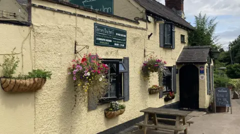 A photo of a pub with yellow walls and a green sign. There are a number of hanging baskets on the wall, a table, door and three windows
