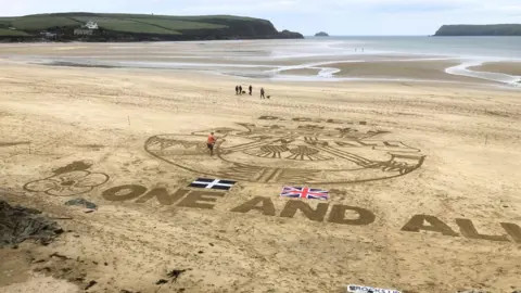 Rocks Up The drawing with the sea extending beyond it and headlands to left and right. One of the men can be seen in the drawing working on the depiction of the horn. There are four people with two dogs walking along the beach.