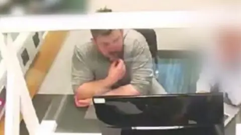 McCullagh in a grey tshirt behind a glass screen looking at a TV screen. 