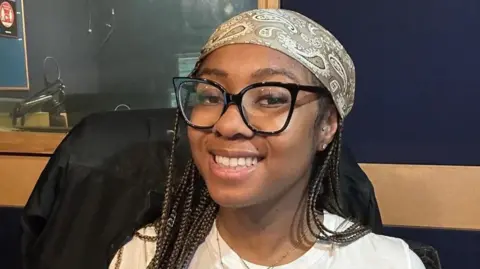 British Jamaican Shaniyaa Holness-Mckenzie smiles at the camera. She wears black rimmed glasses and a bandana.