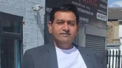 A man with black hair wearing a grey suit over a white T-shirt looks at the camera.