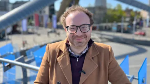 Stephen Richmond has short curly brown hair and wears glasses. He wears a camel skin coat and a red zip up jumper.