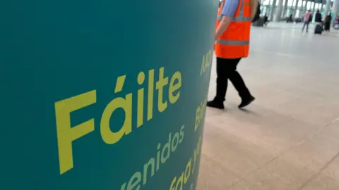 A green sign saying "Fáilte", Irish for "welcome", in the Grand Central Station, with a Translink employee in an orange fluorescent jacket behind. 