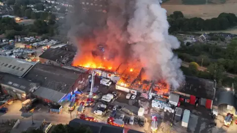 An aerial view of a huge seat of fire across a large industrial complex with a plume of smoke rising high